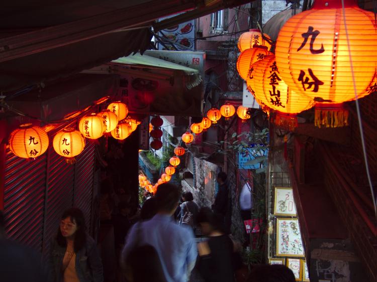 People passing through a narrow alleyway with red lanterns to either side at dusk