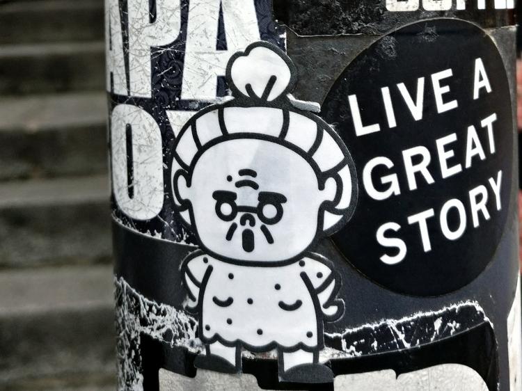Two stickers on a lamppost; one showing a simple cartoon sketch of an old woman, the other a black circle with the words 'live a great story' inside