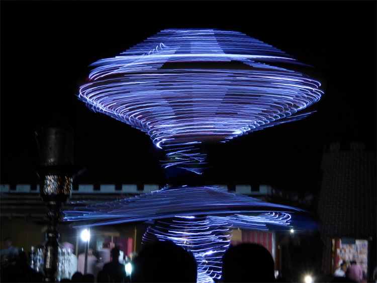 A dancer forming an abstract shape by throwing up parts of their garment with blue lights embedded in it while spinning around