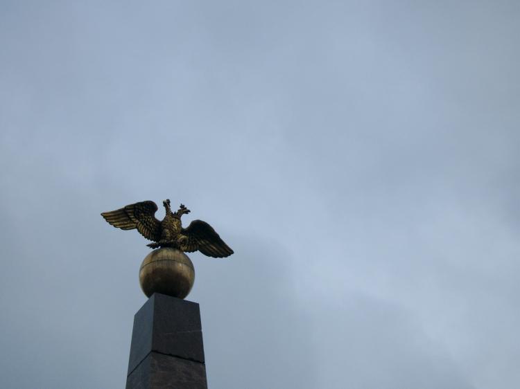A golden sculpture of a double-headed eagle sitting on a gold ball on top of a stone pillar against a cloudy grey sky
