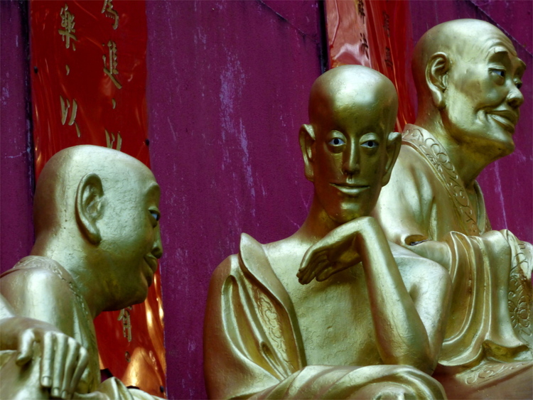 Golden statues of three Buddhist monks, one looking inquisitively at the camera and touching his chin