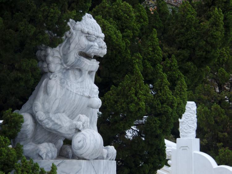 A white marble statue of a lion sitting in a hedge, holding a stone ball with one paw