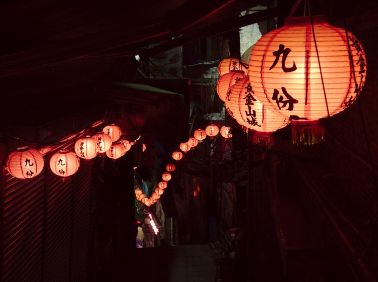 A dark, narrow alleyway of closed shops lined with glowing red lanterns to both side