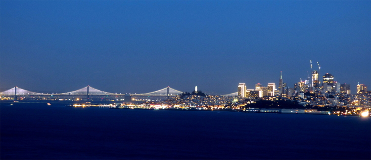 A lit-up cityscape in the distance across the sea, with a long white suspension bridge spanning the water