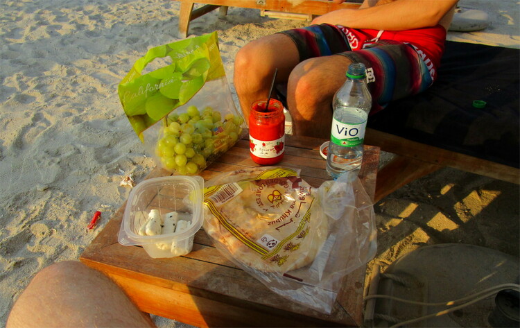 Bags of grapes, bread and halloumi cheese, some water and aivar on a square wooden table on the beach