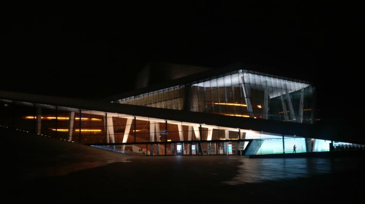 The Oslo Opera House at night. The building's large window-fronts form geometric shapes of orange and blue light against a pitch black sky. A person's silhouette can be seen walking behind one window. 