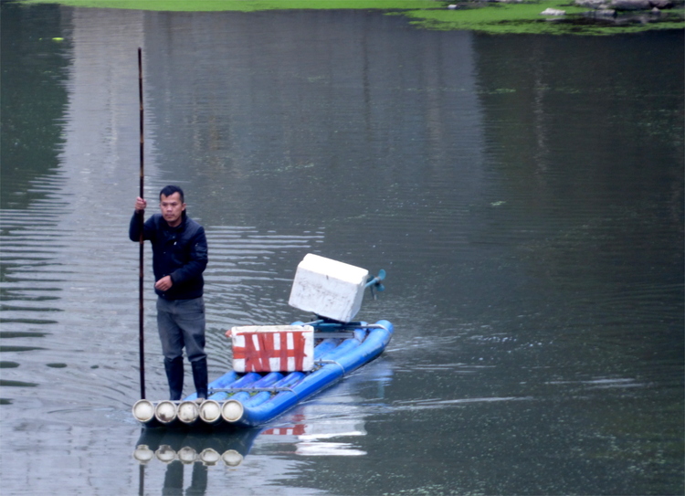 A man holding a long wooden pole standing on a narrow raft made of blue plastic pipes on a river, apparently transporting a white box