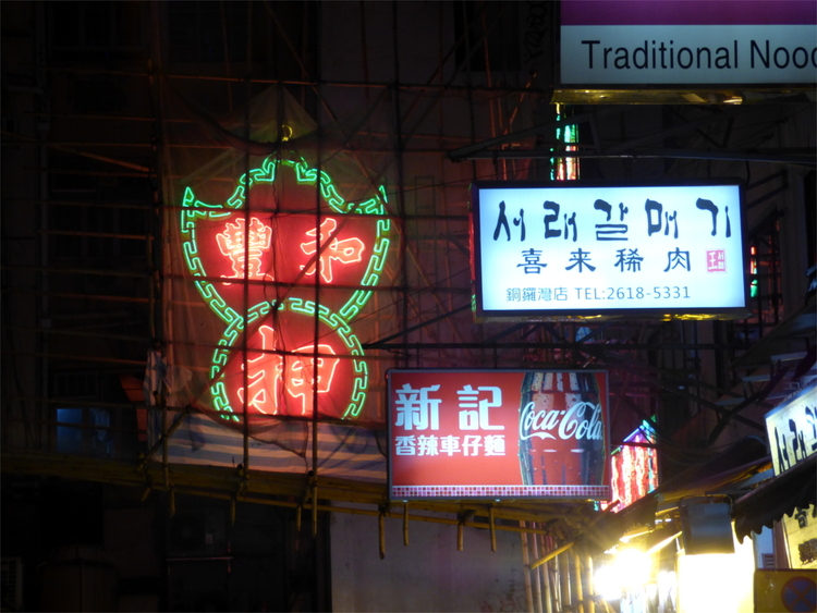 A neon sign showing red Chinese characters in an ornamental green border encased by bamboo scaffolding above a street