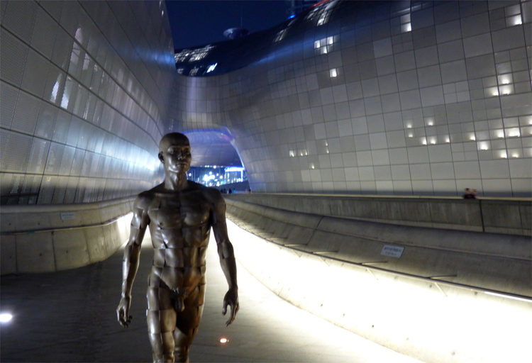 Metal statue of a nude man walking purposefully towards the camera with the organic-shaped buildings of Dongdaemun Design Plaza in the background