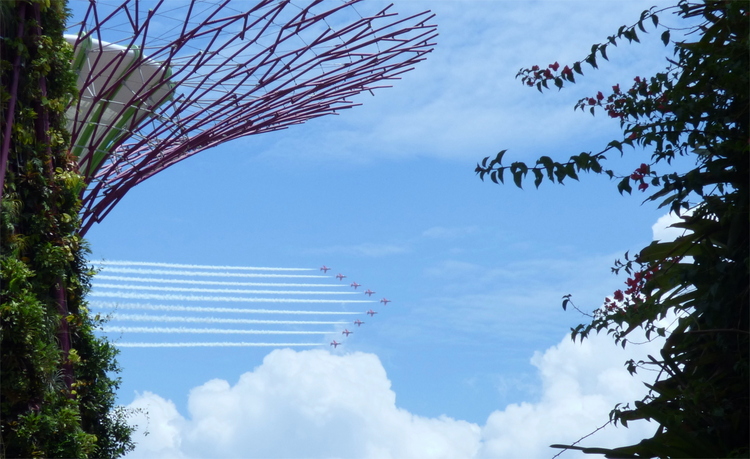 An arrowhead-shaped formation of fighter jets leaving white trails across the sky next to an artificial tree structure