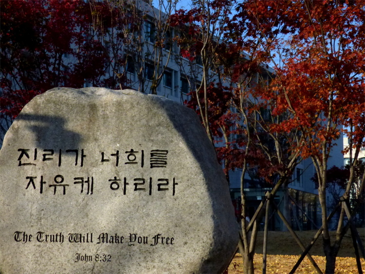 A large rock in front of red-leaved trees bearing a Korean inscription with the English translation 'The Truth Will Make You Free' underneath