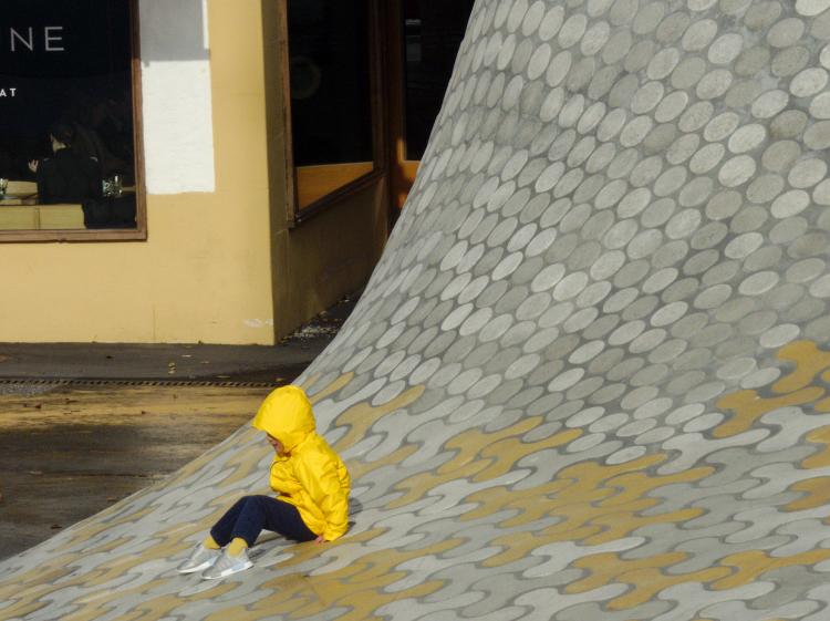 A child in a yellow raincoat sitting on a warped, patterned concrete surface