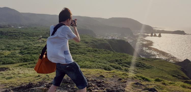 Nils standing on a cliff by the sea with a camera, strong wind blowing in his hair, shirt, and bag
