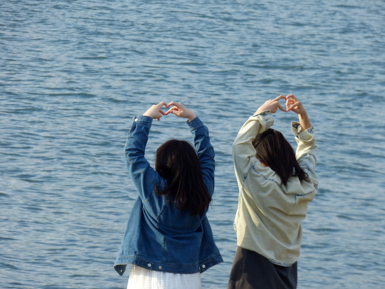 Two women standing at the shore forming heart-shapes with their hands above the heads, posing for a picture with their backs to the camera