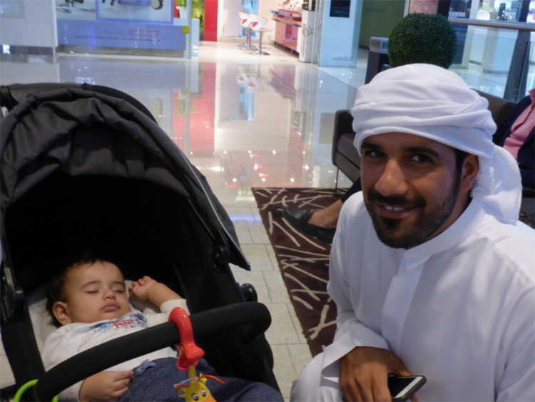 An Arab man in traditional white clothing  kneeling next to his baby son in a stroller