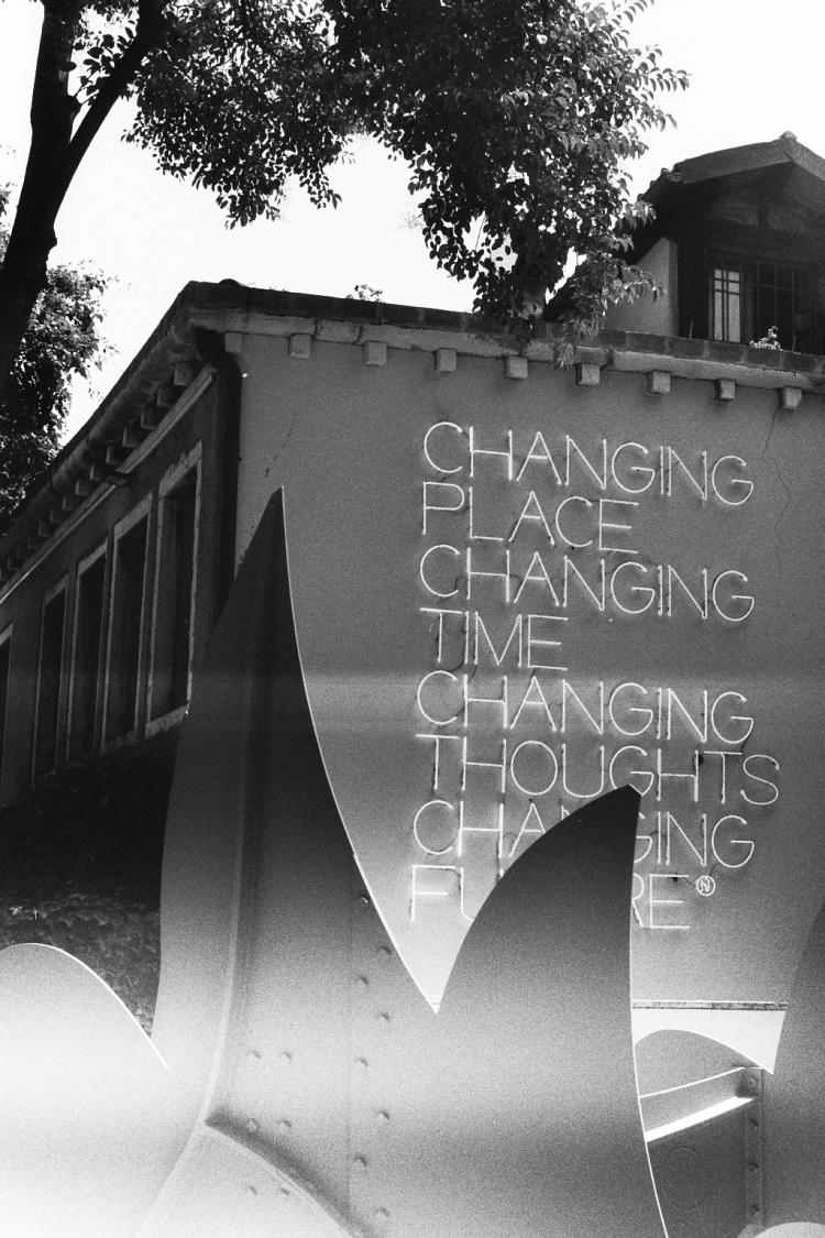 Neon letters reading "changing place / changing time / changing thoughts / changing future" on a wall behind an abstract steel sculpture