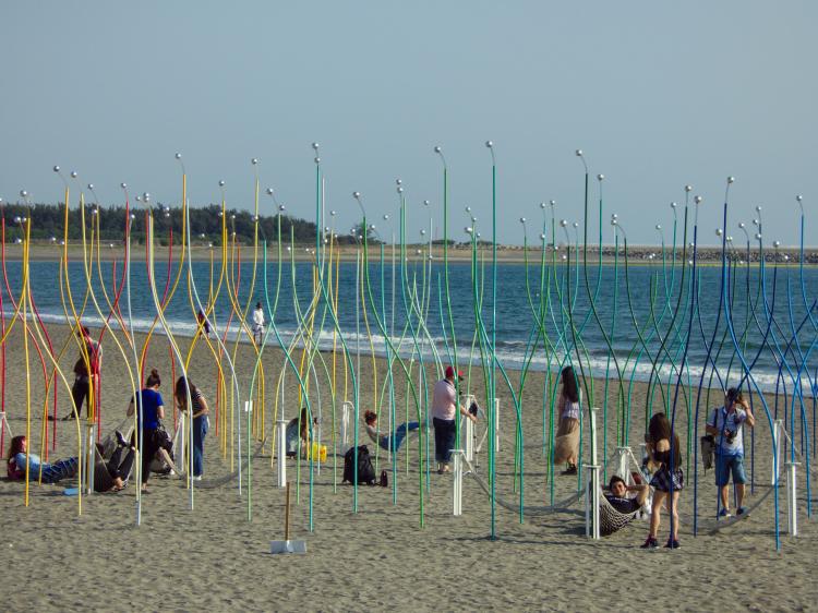 A beach area with bent metal poles in rainbow-shades sticking out from the sand, some hammocks suspended between them with people resting or taking pictures in the installation