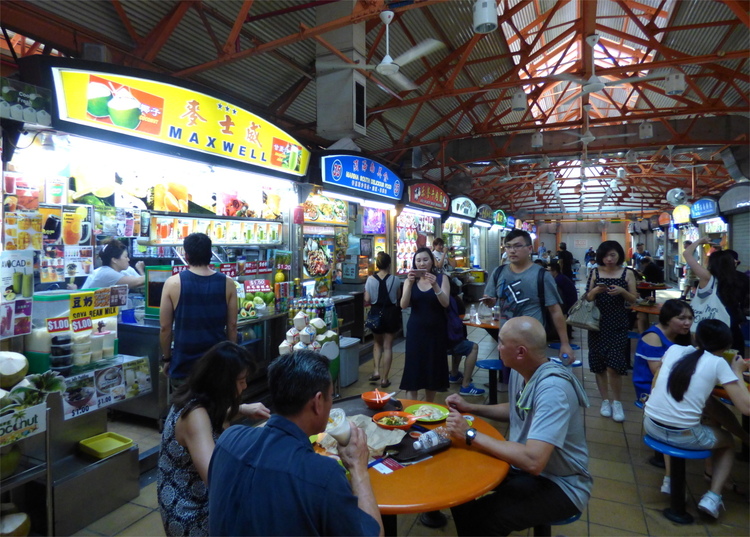 A busy indoor food court with many small food vendors and customers eating together on round tables