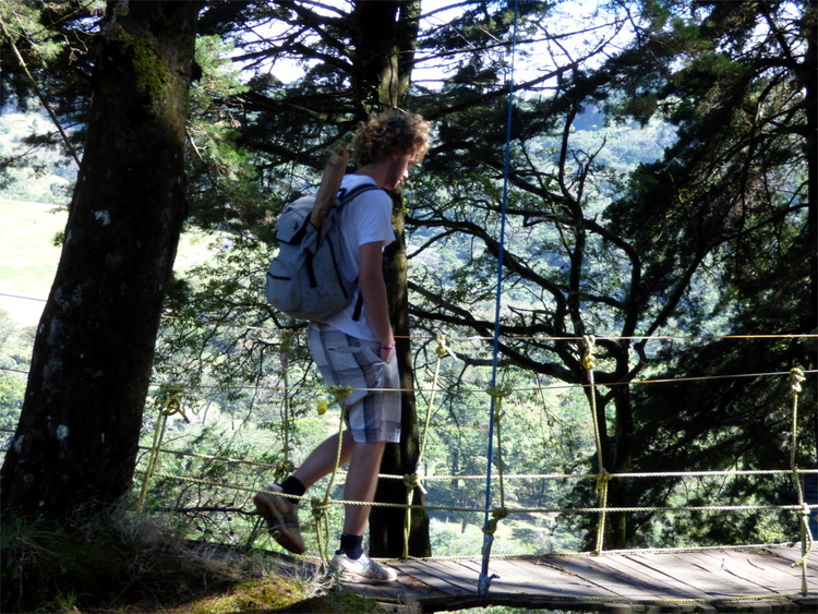 Willi wearing shorts and a T-Shirt, walking across a small wooden hang-bridge carrying a baguette in his backpack 