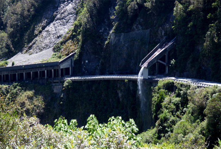 A narrow mountain road emerging from a tunnel and passing underneath a concrete spillway diverting a small stream above it