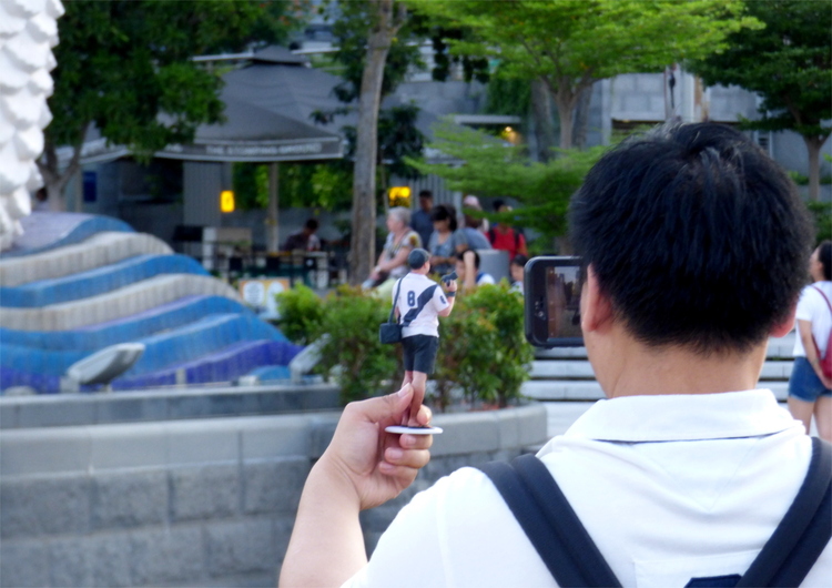 A man taking a picture of a 3D-printed miniature of himself, wearing the same black-and-white sports shirt