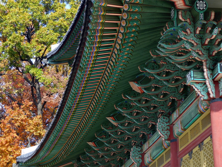 The intricate green and red wooden roof construction of a Korean palace with matching autumn foliage in the background