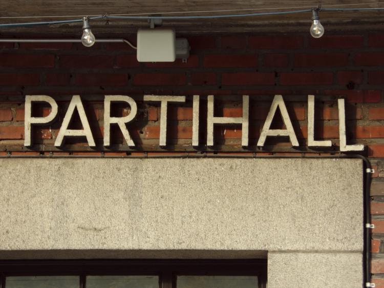 Capital letters spelling the word 'Partihall' above a door frame on a red brick wall