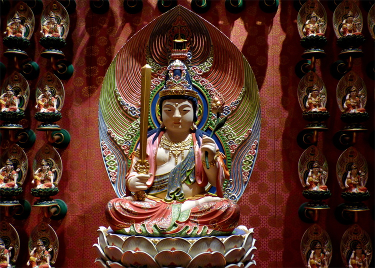 A colourful Buddha statue sitting in a lotus flower holding a stylized sword, with many smaller statues on the wall around it
