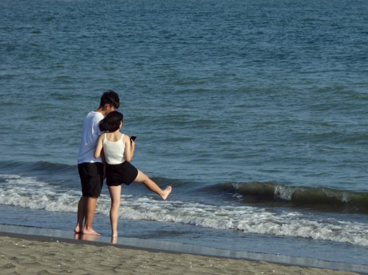 A young couple in matching white tops and black shorts standing next to each other on the shoreline, the woman stretching out one leg towards the water