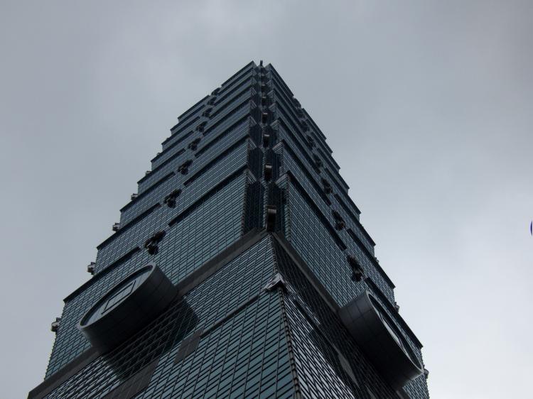 A look up from the base of the Taipei 101, looking like a geometric bamboo plant made of dark blue and grey glass against a cloudy sky