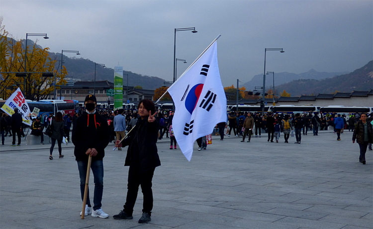 Two young men in black standing on a square with a crowd in the background, one of them holding a South Korean flag and making the 'peace' sign at the camera