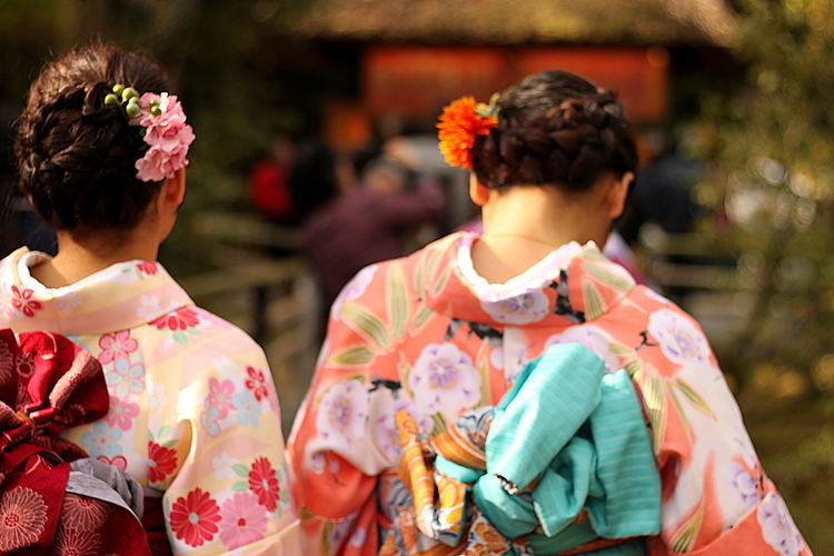 Two women in elaborate traditional Japanese clothing with flowers braided into their hair walking away from the camera towards a temple