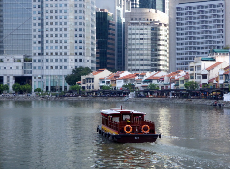 A river boat making its way down a body of water bordered by a row of red-roofed houses with skyscrapers in the background