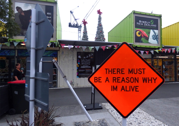 A diamond-shaped neon orange street-sign reading 'there must be a reason why im alive' in front of some shipping container buildings
