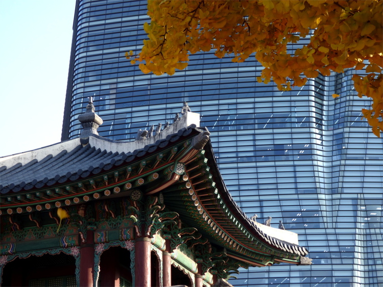 An intricate, colourful wooden roof of a Korean palace in front of a modern skyscraper's glass facade and bright yellow ginkgo leaves