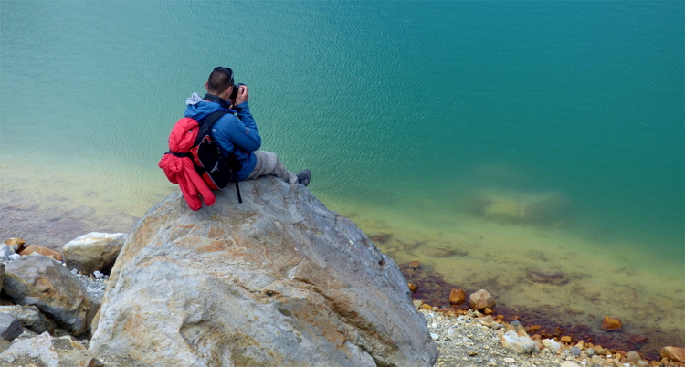 A man in hiking gear sitting on a large stone photographing a colourful green, red and yellow volcanic lake