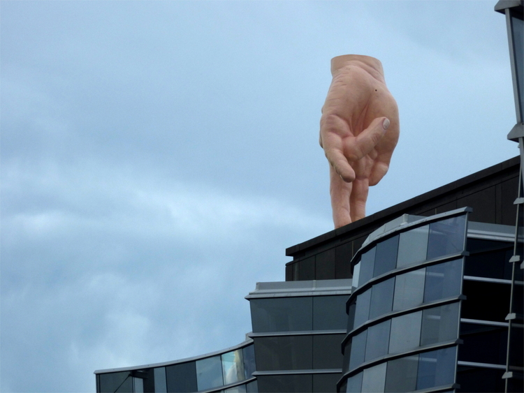 A large, realistic sculpture of a hand walking on two fingers on the roof of a futuristic glass building