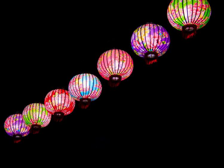 A string of colourful lit lanterns with pink floral motives against a black background