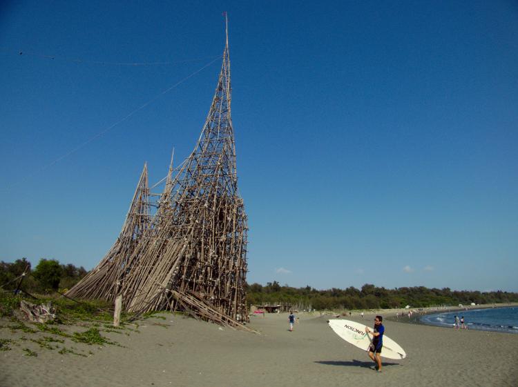 A surfer carrying their board across the beach with a large castle-like construction made of wooden stakes behind them