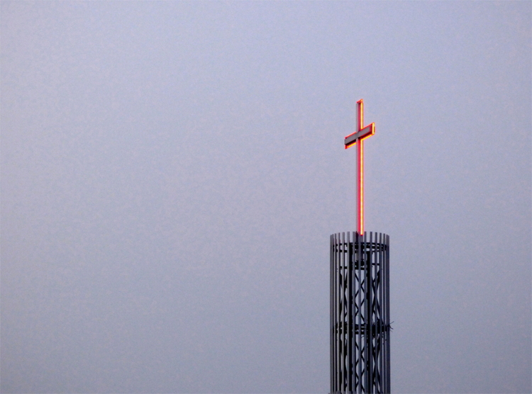 A Christian neon cross glowing red on a metal tower against a grey sky