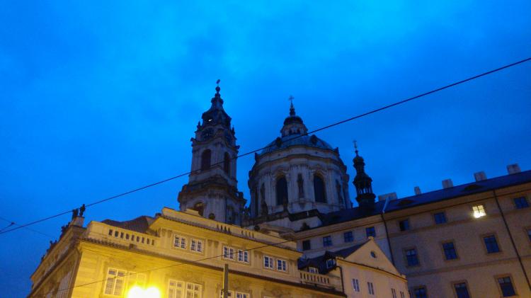 A baroque church rising up from behind a row of residential buildings against a deep blue evening sky
