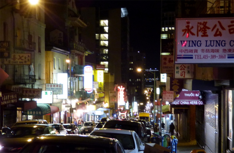 View of a street with parked cars at night, with many signs featuring Chinese characters advertising various businesses