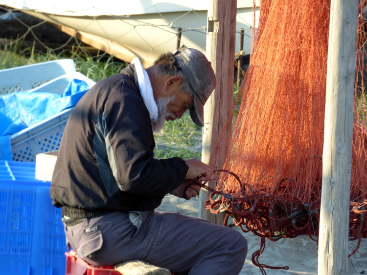 A bearded old fisherman wearing a baseball cap sitting on a stool repairing an orange fishing net