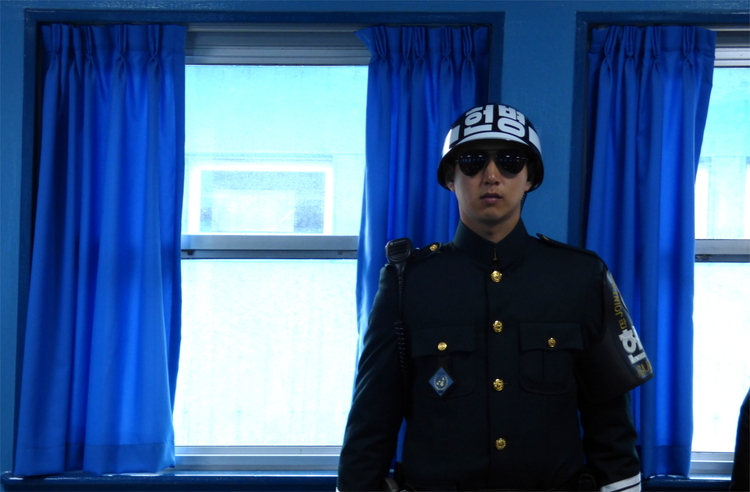 A stern-looking South Korean soldier in uniform with dark sunglasses and a metal helmet standing in front of a blue wall with two windows and blue curtains