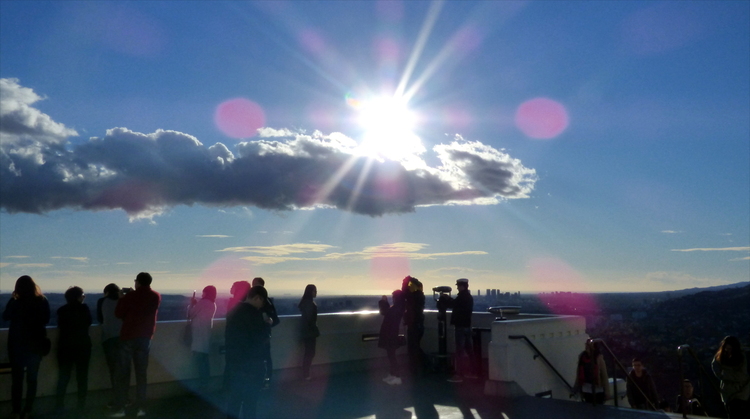 People standing on a railing looking into the distance with the glaring sun low in the sky above a single cloud