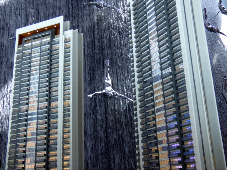 The sculpture of a human diving down a waterfall seen through the space between two miniature models of skyscrapers, appearing to touch both sides of them in free fall
