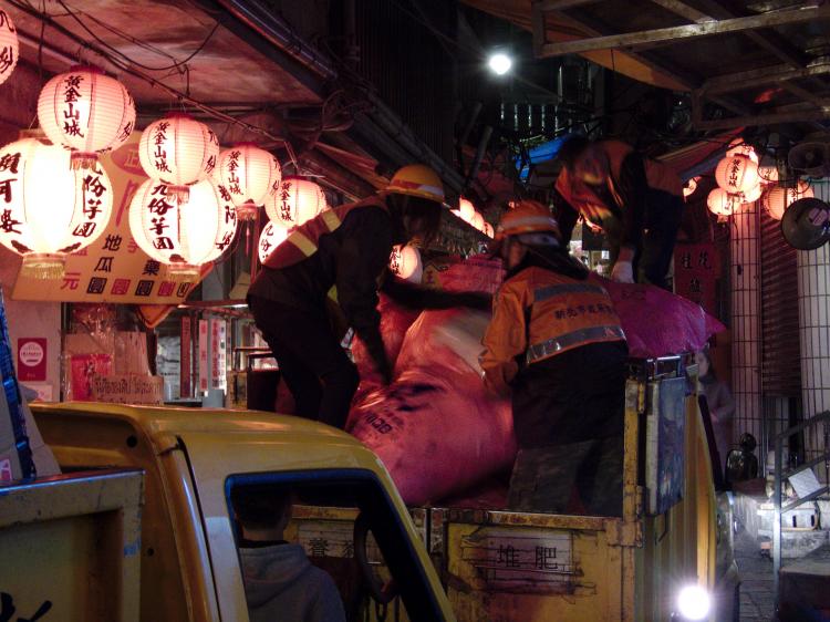 Garbage-men loading bags onto tiny garbage trucks passing through a narrow street lit by red lanterns