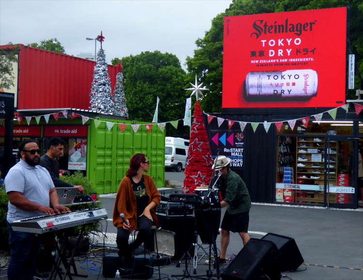 A street-music band of four people performing on a public square enclosed by shipping container buildings with a red advertisement screen for Tokyo Dry beer