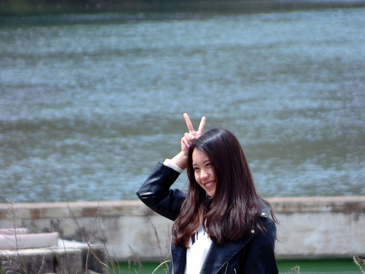 A woman smiling and posing for pictures in front of a river, making a bunny-ear gesture behind her own head