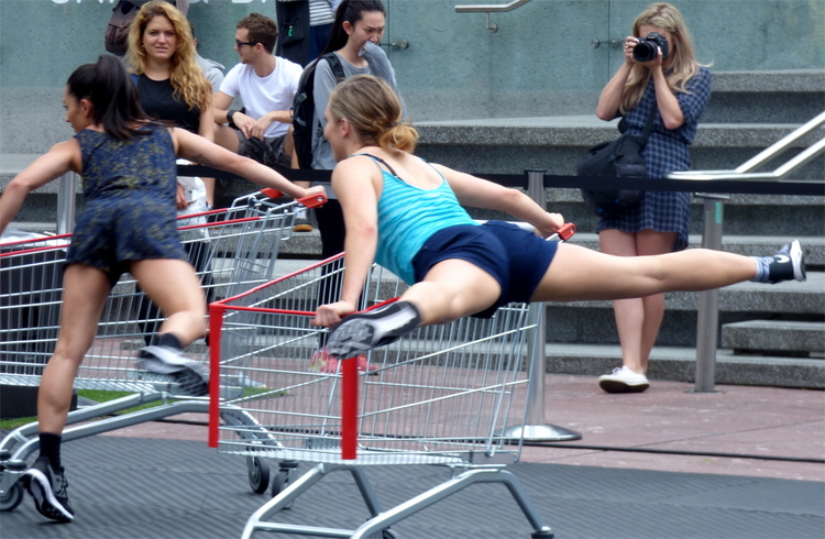 Two women performing acrobatics on shopping carts, holding on to the side of the cart while lifting their legs up and spinning around in the air